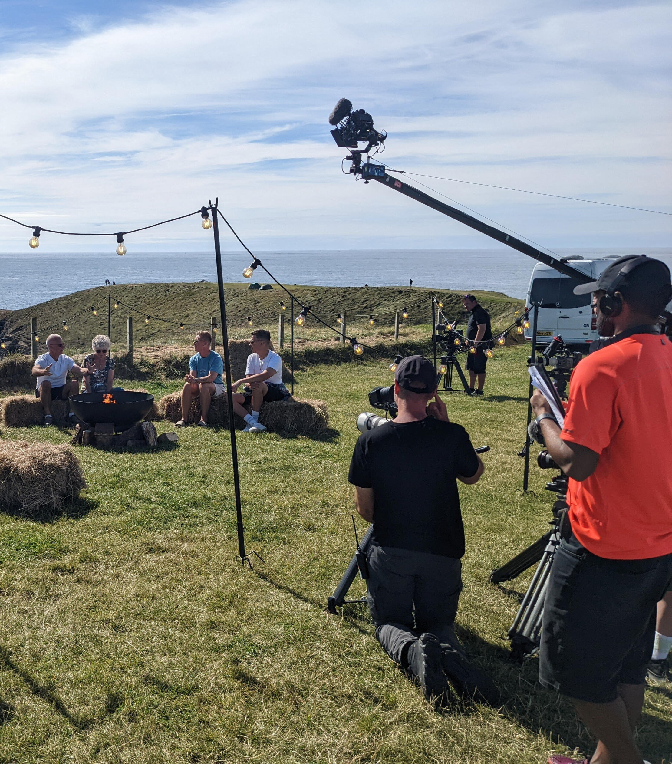 A camera crew filming influencers on a campsite on a grassy hill overlooking the ocean on a sunny day.